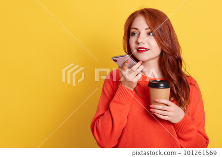 Attractive red haired woman recording voice messages, holding cell phone and coffee to go in hands, looking away, copy space for advertisement, isolated over yellow background. Attractive red haired woman recording voice messages, holding cell phone and coffee to go in hands, looking away, copy space for advertisement, isolated over yellow background. 101261569