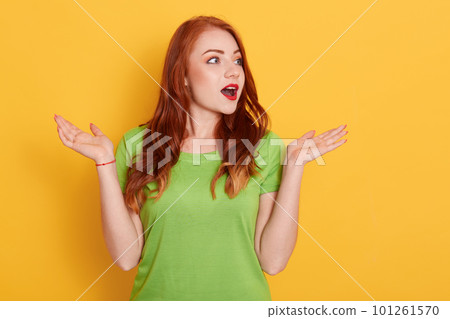 Close up portrait of beautiful red haired woman spreading hands and looking aside, standing isolated over yellow background, astonished female with widely opened mouth. Close up portrait of beautiful red haired woman spreading hands and looking aside, standing isolated over yellow background, astonished female with widely opened mouth. 101261570