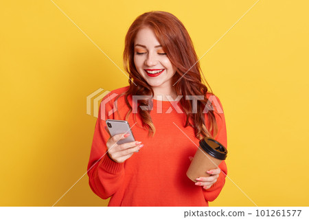 Glad female student has coffee break after lectures, enjoys free time, uses mobile phone for chatting online, looking at device, holding disposable cup with hot beverage, stands against yellow wall. Glad female student has coffee break after lectures, enjoys free time, uses mobile phone for chatting online, looking at device, holding disposable cup with hot beverage, stands against yellow wall. 101261577