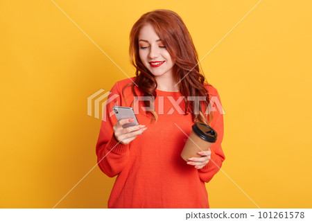 Adorable girl wearing orange sweater posing isolated over yellow background, holding coffee to take away and mobile phone, looks at device's screen with smile. 101261578