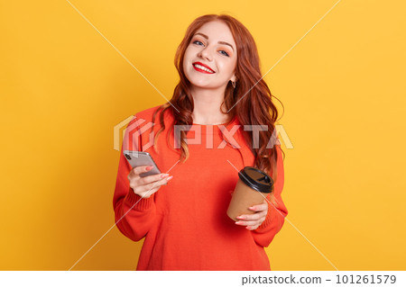 Pleased European woman dressed in casual orange sweater, poses over yellow background, looks smiling at camera, holding smart phone and coffee to go. 101261579