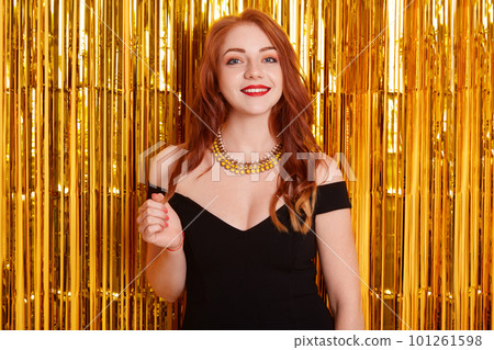Young gorgeous elegant sensual red haired European woman wearing fashion black dress standing against wall decorated with golden tinsel, happy female with red lips wears black dress, being at party. Young gorgeous elegant sensual red haired European woman wearing fashion black dress standing against wall decorated with golden tinsel, happy female with red lips wears black dress, being at party. 101261598