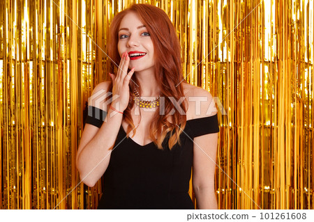 Surprised lady with beautiful red hair, wearing black elegant dress with bare shoulders and necklace, looking at camera and covering her mouth, celebrating her birthday. Surprised lady with beautiful red hair, wearing black elegant dress with bare shoulders and necklace, looking at camera and covering her mouth, celebrating her birthday. 101261608