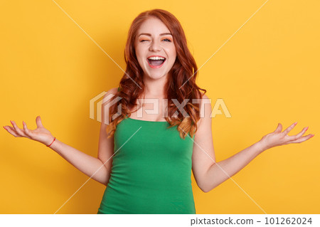 Happy exited woman posing with spreading hands aside and blinking eyes, wearing green outfit, posing isolated over yellow background, lady looks directly at camera. Happy exited woman posing with spreading hands aside and blinking eyes, wearing green outfit, posing isolated over yellow background, lady looks directly at camera. 101262024