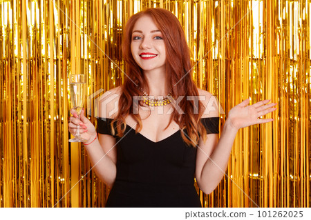 Portrait of red haired beautiful girl with smile on her face, bright make-up and hairstyle, celebrating holiday, holding glass of wine in her hands. 101262025