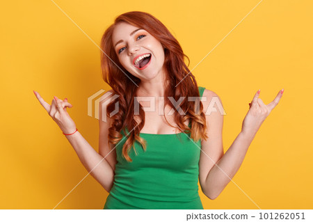 Young redhaired woman isolated over yellow background showing rock gesture with fingers, keeps mouth opened, dresses green outfit, lady being at party. Young redhaired woman isolated over yellow background showing rock gesture with fingers, keeps mouth opened, dresses green outfit, lady being at party. 101262051
