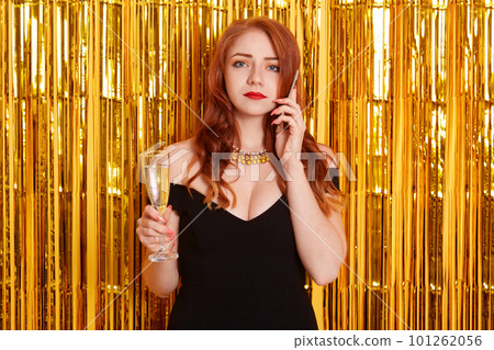 Upset red haired female talking via phone, hears unpleasant news, holding glass of wine in hands, standing against bright sparkle wall decorated with tinsel, lady wearing black dress. 101262056