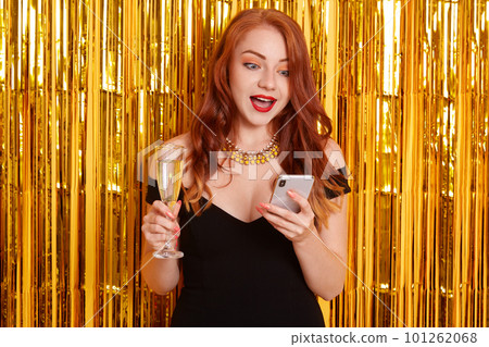 Astonished female with red lips and widely opened mouth, looking with shocked facial expression at her device, girl wearing black dress, posing isolated over wall decorated with golden tinsel. 101262068