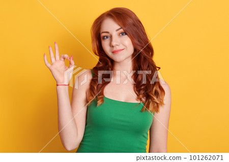 Calm red haired young woman shows okay sign with hand, demonstrates approval, wears green clothing, isolated over yellow background, female looks at camera with smile. Calm red haired young woman shows okay sign with hand, demonstrates approval, wears green clothing, isolated over yellow background, female looks at camera with smile. 101262071
