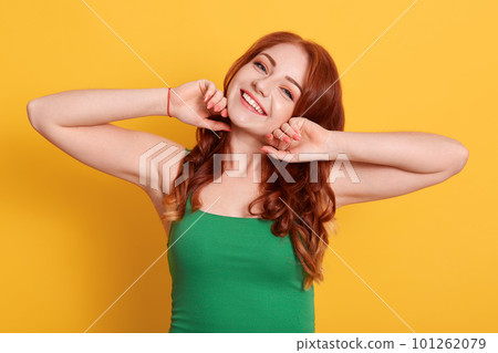 Photo of greatly red haired female wearing green casual t shirt, indicates at yellow wall, looking smiling directly at camera, keeping hands on her face. Photo of greatly red haired female wearing green casual t shirt, indicates at yellow wall, looking smiling directly at camera, keeping hands on her face. 101262079