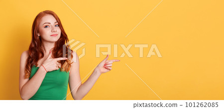 Smiling Caucasian woman with curls, wears green t shirt, points aside with index fingers, isolated over yellow background, copy space for promotional text. Smiling Caucasian woman with curls, wears green t shirt, points aside with index fingers, isolated over yellow background, copy space for promotional text. 101262085