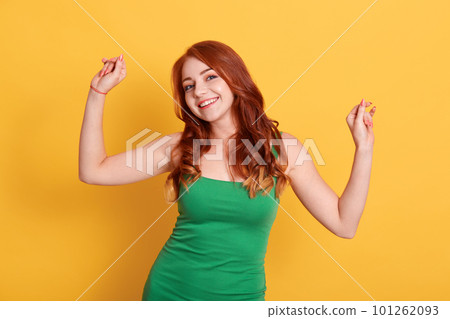 Optimistic red haired young woman poses with hands up, dances carefree, wears green sleeveless t shirt, looks at camera, isolated over yellow wall, copy space for your promotion. 101262093