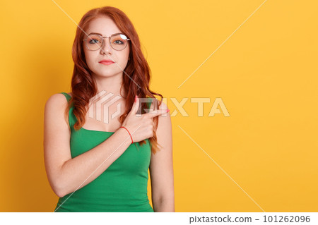 Pleasant looking woman in eyewear points with index finger at free space, models against yellow background, dressed green clothing, european young female demonstrates something aside. 101262096