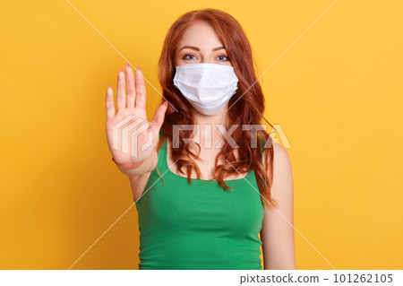 Serious redhaired woman makes stop gesture, pulls palm towards camera, wears medical flu mask and green shirt, stay at home not to spread coronavirus disease. 101262105