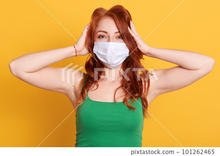 Woman in medical mask plugging ears with hands. European young redhead female ignoring noise or news about coronavirus, wearing green t shirt, posing against yellow wall. Woman in medical mask plugging ears with hands. European young redhead female ignoring noise or news about coronavirus, wearing green t shirt, posing against yellow wall. 101262465