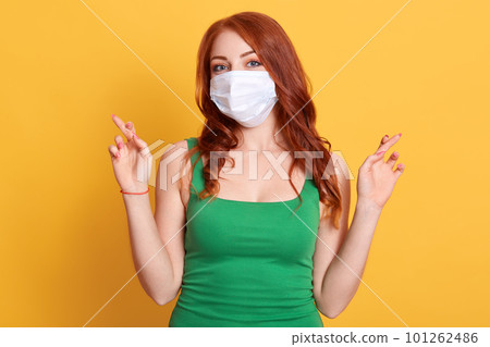 Red haired young female with crossed fingers, wishing best, standing against yellow background, ginger woman wearing medical mask, wants to avoid virus. 101262486