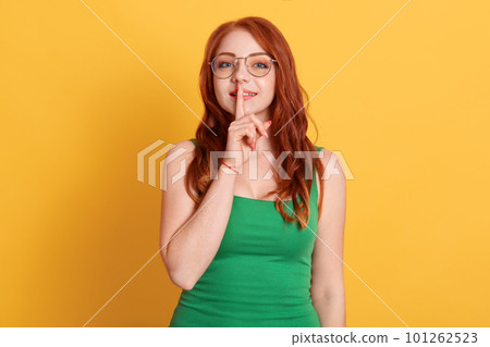 Closeup photo of attractive pretty cute nice glad charming lady holding forefinger near mouth isolated over yellow background, red haired female in green t shirt and spectacles. 101262523