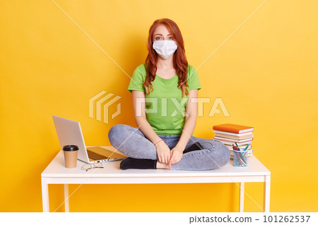 Photo of smart intelligent student girl sits on table, wearing medical mask, isolated over yellow background, lady with red hair keeps crossed legs, preparing for classes, posing near laptop and books Photo of smart intelligent student girl sits on table, wearing medical mask, isolated over yellow background, lady with red hair keeps crossed legs, preparing for classes, posing near laptop and books 101262537
