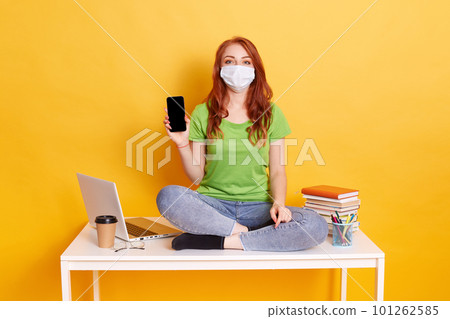 Red haired woman studies while sitting on desk with laptop and books isolated on yellow background. Distance education at home. Lady holding mobile phone with blank empty screen. Red haired woman studies while sitting on desk with laptop and books isolated on yellow background. Distance education at home. Lady holding mobile phone with blank empty screen. 101262585