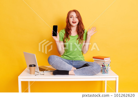 Young European woman showing smart phone sitting on table with crossed legs, wearing green t shirt and jeans, showing phone with blank screen and raising hand, has surprised facial expression. 101262620