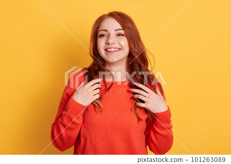 Cheerful gorgeous young woman wearing orange jumper, smiling happily while looking aside. Pretty girl excited joyful smile touching her red hair against yellow background. 101263089