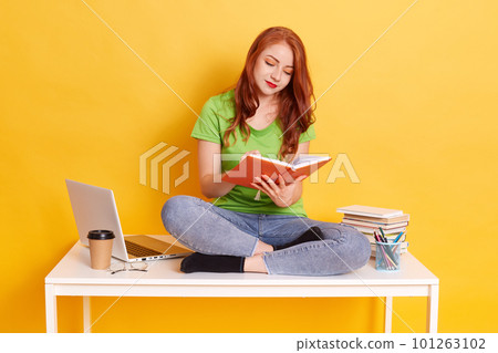 Student girl sitting on desk with crossed legs and reading book, red haired female wearing green casual t shirt and jeans, woman studying against yellow wall surrounded with laptop computer, coffee. 101263102