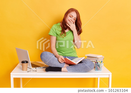 Adorable red haired tired sleepy woman sitting on her desk with books and computer, keeps eyes closed, covering her mouth while yawning, spending long hours studying against yellow wall. Adorable red haired tired sleepy woman sitting on her desk with books and computer, keeps eyes closed, covering her mouth while yawning, spending long hours studying against yellow wall. 101263104