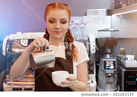 Red haired barista girl brews coffee from turk in coffee shop, concentrated woman working in cafe, lady in white t shirt and brown apron with coffee machine in background. Red haired barista girl brews coffee from turk in coffee shop, concentrated woman working in cafe, lady in white t shirt and brown apron with coffee machine in background. 101263137
