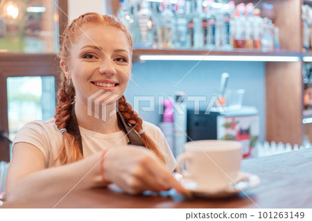 Young smiling woman standing at bar in coffee shop, barista posing at counter, offers cup of hot beverage for her client, red haired female working in cafe. Young smiling woman standing at bar in coffee shop, barista posing at counter, offers cup of hot beverage for her client, red haired female working in cafe. 101263149