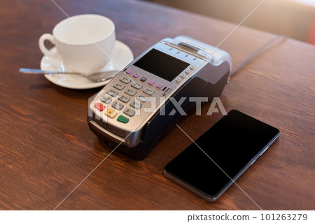 Paying for coffee, white ceramic cup, terminal and phone with blank screen on brown counter, device for contactless payment in coffee shop. Paying for coffee, white ceramic cup, terminal and phone with blank screen on brown counter, device for contactless payment in coffee shop. 101263279