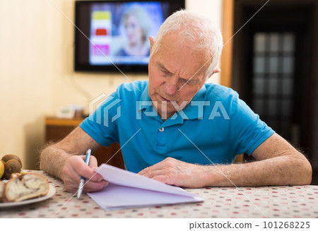 Serious older man writing letter at table in living room Serious older man writing letter at table in living room 101268225