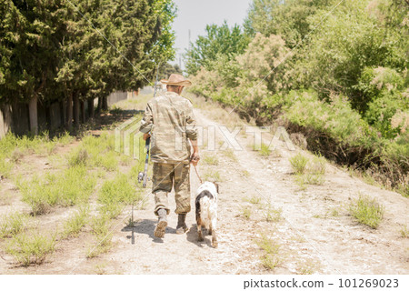 Unrecognizable, slender male fisherman in camo outfit walking down dirt road with obedient spotted dog in summer. Back view of adult guy with fishing pole going along bushy path. Concept of hobby. Unrecognizable, slender male fisherman in camo outfit walking down dirt road with obedient spotted dog in summer. Back view of adult guy with fishing pole going along bushy path. Concept of hobby. 101269023