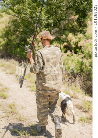 Unrecognizable fisherman in camouflage going on fishing with setter dog at daytime. Back view of slim fisher carrying big rod, while walking along path to lake outdoors. Concept of hobby, lifestyle. 101269025