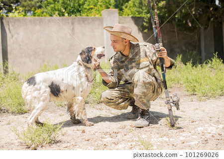 Caring fisherman with fishing pole, petting obedient dog, while sitting on tiptoe outdoors. Side view of male fisher and pet looking to each other with love, while resting. Concept of animal care. 101269026