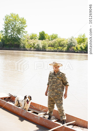 Concentrated male angler with faithful dog standing in old boat, going mooring after fishing in countryside. Front view of male fisherman enjoying trip on murky river. Concept of fishery, hobby. Concentrated male angler with faithful dog standing in old boat, going mooring after fishing in countryside. Front view of male fisherman enjoying trip on murky river. Concept of fishery, hobby. 101269046