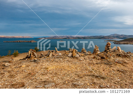 Stones piled by pyramids and a view of Lake Baikal. Hills beyond the lake. Several islands in the lake. Cloudy and gloomy. Horizontal. Stones piled by pyramids and a view of Lake Baikal. Hills beyond the lake. Several islands in the lake. Cloudy and gloomy. Horizontal. 101271169