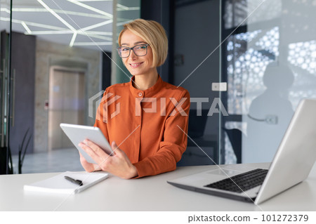 Portrait of a young female psychologist, psychotherapist, tutor sitting in the office at a table with a laptop, holding a tablet in her hands and looking at the camera smiling. Portrait of a young female psychologist, psychotherapist, tutor sitting in the office at a table with a laptop, holding a tablet in her hands and looking at the camera smiling. 101272379