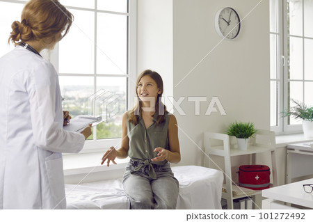 Smiling teenage girl talks about her health to female doctor during appointment in medical office. 101272492