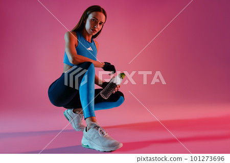 Healthy woman sitting with water bottle in sport clothing over studio background Healthy woman sitting with water bottle in sport clothing over studio background 101273696