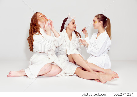Three beautiful young girls in bathrobes applying face masks, taking care after skin against white background Three beautiful young girls in bathrobes applying face masks, taking care after skin against white background 101274234