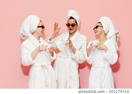 Three cheerful pretty young girls in bathrobes and towels, drinking coffee after spa against pink studio background Three cheerful pretty young girls in bathrobes and towels, drinking coffee after spa against pink studio background 101274318