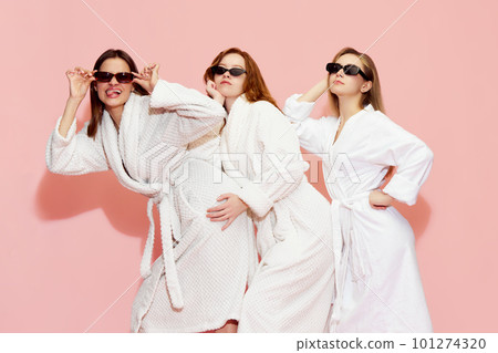 Three attractive young girls in white bathrobe and sunglasses posing against pink studio background. Sleepover 101274320