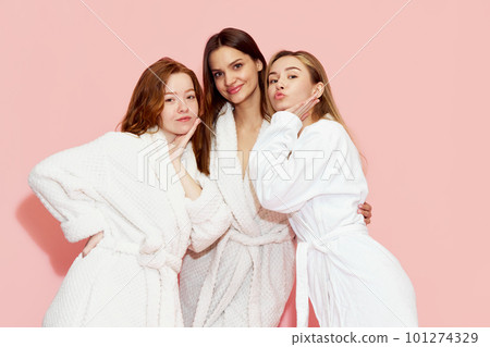 Portrait od three beautiful girls, friends in white bathrobes posing against pink studio background. Spa weekends, self-care 101274329