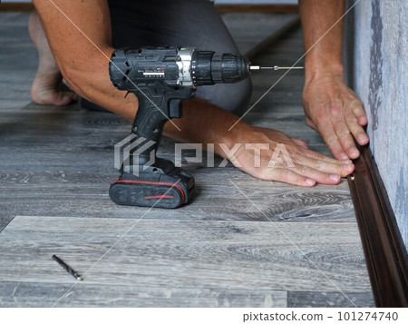 a man installs a floor wooden plinth made of dark lacquered wood in a room with gray textured linoleum using a screwdriver, holding the plank with his hands at the junction of wall and floor a man installs a floor wooden plinth made of dark lacquered wood in a room with gray textured linoleum using a screwdriver, holding the plank with his hands at the junction of wall and floor 101274740