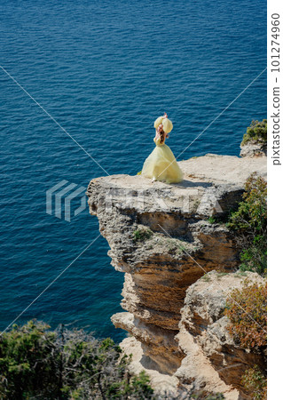 Woman in a yellow dress on the sea. Side view Young beautiful sensual woman in yellow long dress posing on a rock high above the sea at sunset. Girl in nature against the blue sky Woman in a yellow dress on the sea. Side view Young beautiful sensual woman in yellow long dress posing on a rock high above the sea at sunset. Girl in nature against the blue sky 101274960