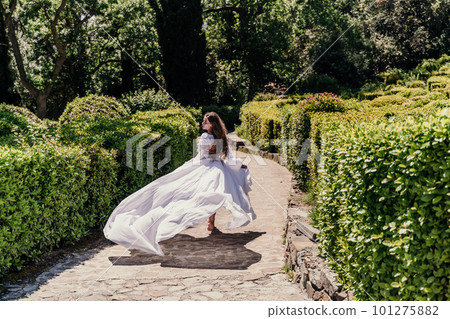 Brunette runs white dress park. A beautiful woman with long brown hair and a long white dress runs along the path along the beautiful bushes in the park, rear view Brunette runs white dress park. A beautiful woman with long brown hair and a long white dress runs along the path along the beautiful bushes in the park, rear view 101275882