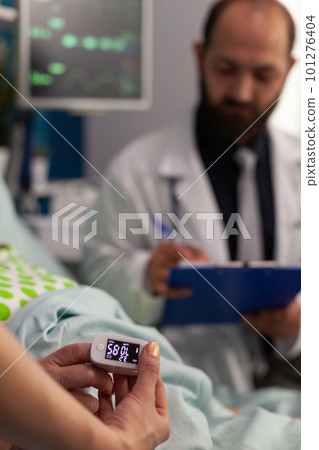 Nurse measuring blood oxygen level with oximeter on elderly patient lying in hospital bed. Male doctor reviewing results of medical examinations of retired woman, specialized care. 101276404