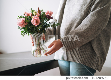 Young woman in grey knitted sweater holding vase with bouqet of flowers at the home 101289730