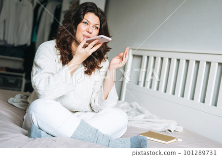 Young adult brunette woman in white knitted sweater using mobile phone on the bed at home Young adult brunette woman in white knitted sweater using mobile phone on the bed at home 101289731