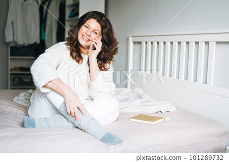 Young adult smiling brunette woman in white knitted sweater using mobile phone on bed at home 101289732